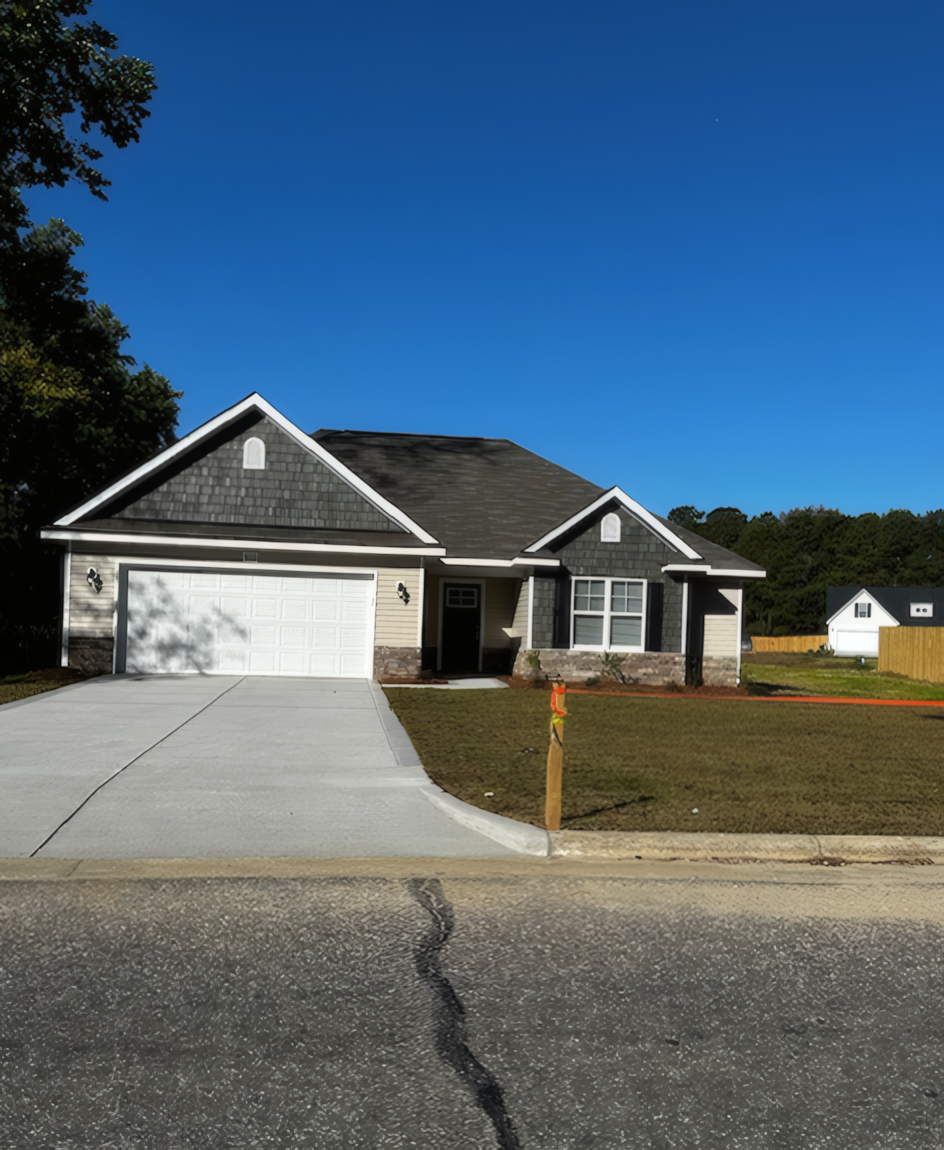 Distance view of dark grey house with large white garage with blue sky above. The view also includes the large cement driveway and road