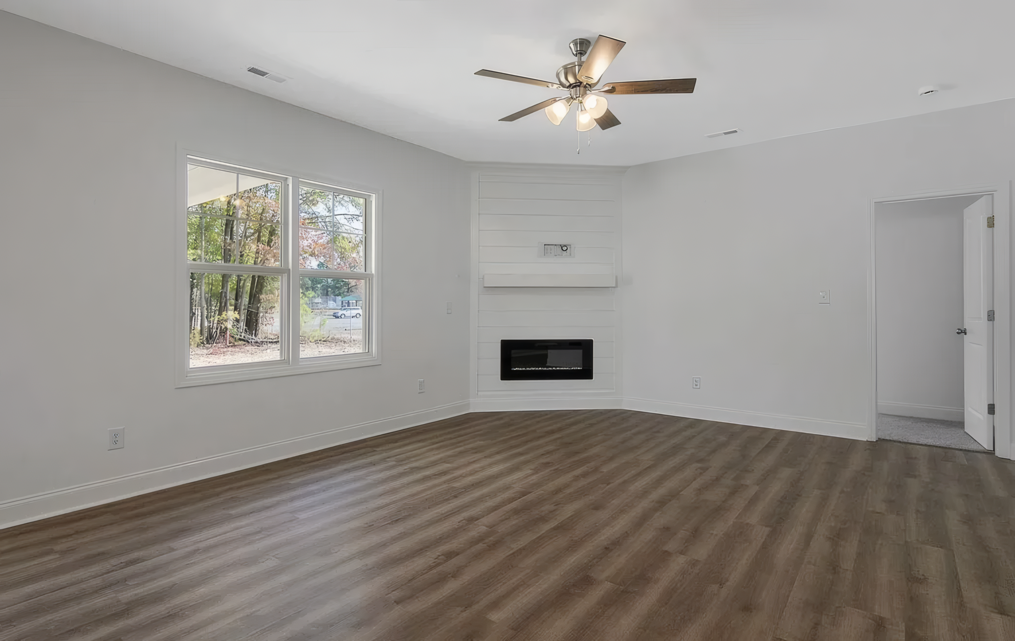 Spacious room with ceiling fan and one giant window. Also has a fireplace and wood pattern floor and white walls